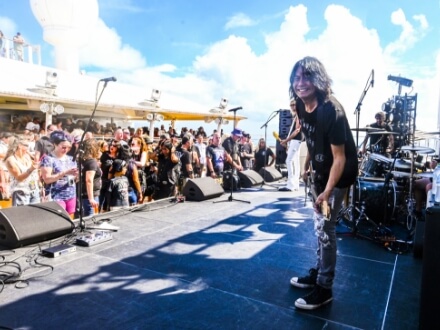 | Monsters Of Rock Cruise Guitarist on stage smiling as a crowd watches a live outdoor concert on a sunny day.