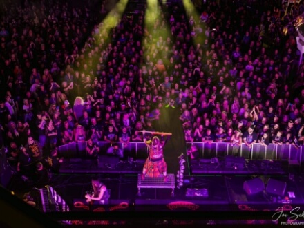 10 | Monsters Of Rock Cruise Energetic live concert scene: center-stage performer in red outfit raises arms, surrounded by a packed audience under purple and yellow lights.