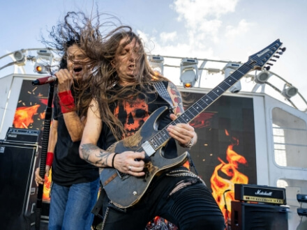 4 | Monsters Of Rock Cruise Two rock guitarists perform on an outdoor stage with a flaming background on a large screen behind them.