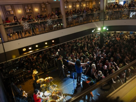 7 | Monsters Of Rock Cruise Band performing on stage in a mall atrium, surrounded by a large audience on two levels