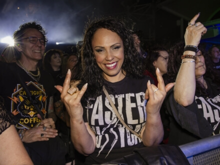 8 | Monsters Of Rock Cruise Smiling woman at a concert barrier, flashing rock hand signs with both hands as crowds cheer behind her.