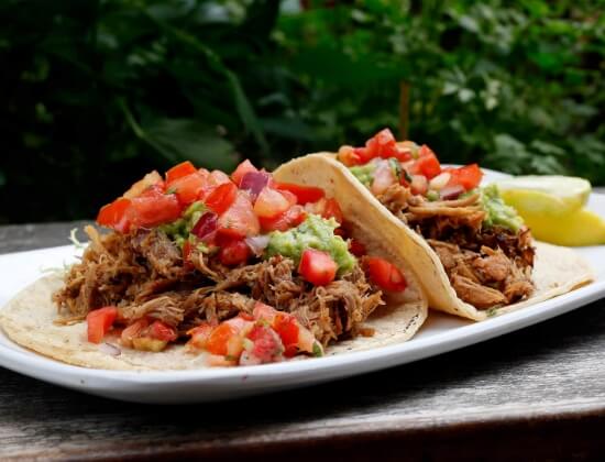 Two soft tacos filled with shredded beef, pico de gallo, and guacamole on a white plate outdoors with a lime wedge nearby