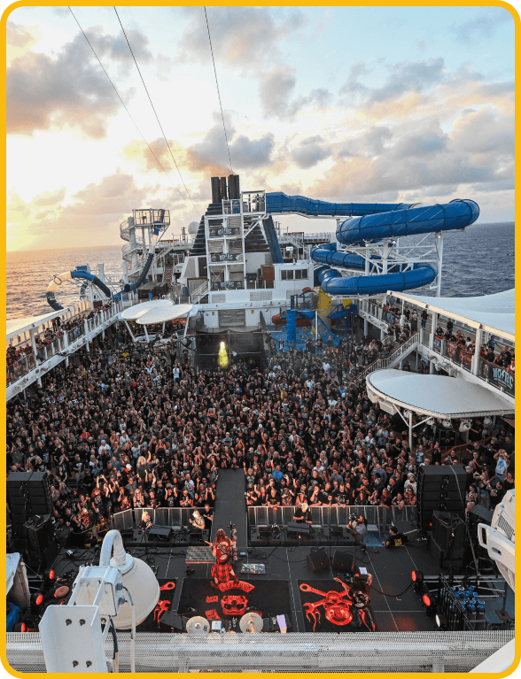 | Monsters Of Rock Cruise Crowd watching a live concert on the deck of a cruise ship at sunset, with stage lights and equipment visible.