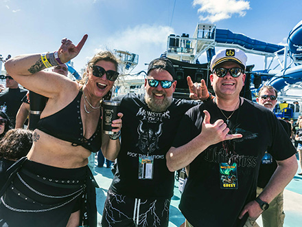 2 | Monsters Of Rock Cruise Group of three friends posing and cheering on a sunny outdoor deck, wearing festival gear and sunglasses, with a water slide in the background.