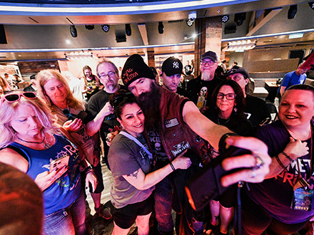 slider 2 | Monsters Of Rock Cruise A diverse group of people posing for a selfie with a smartphone, many wearing badges, in a well-lit backstage area/events setting.